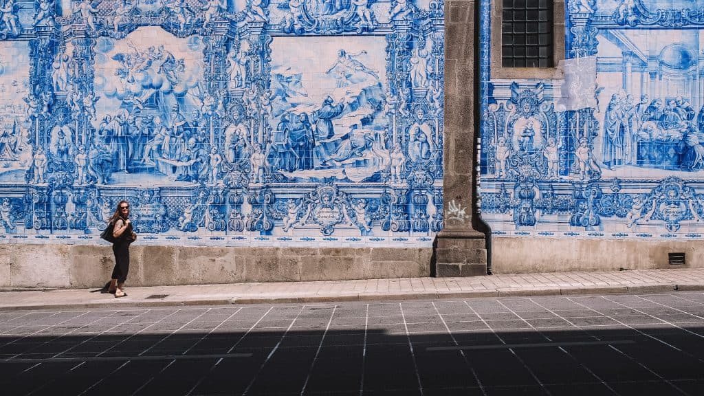 The azulejo-tiled façade of Igreja do Carmo in Porto, Portugal, featuring intricate blue and white ceramic murals depicting historical and religious scenes. A woman in black walks past the ornate tilework, which is framed by dark stone columns. The church's iconic azulejos, added in the early 20th century, highlight Portugal's rich artistic heritage.