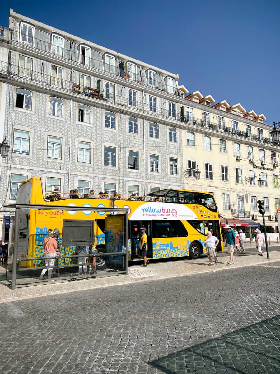 A bright yellow hop-on hop-off bus tour Lisbon vehicle stops at a bustling square with traditional tiled buildings in the background. Tourists board the open-top bus, enjoying the opportunity to explore Lisbon's iconic attractions on a sunny day.