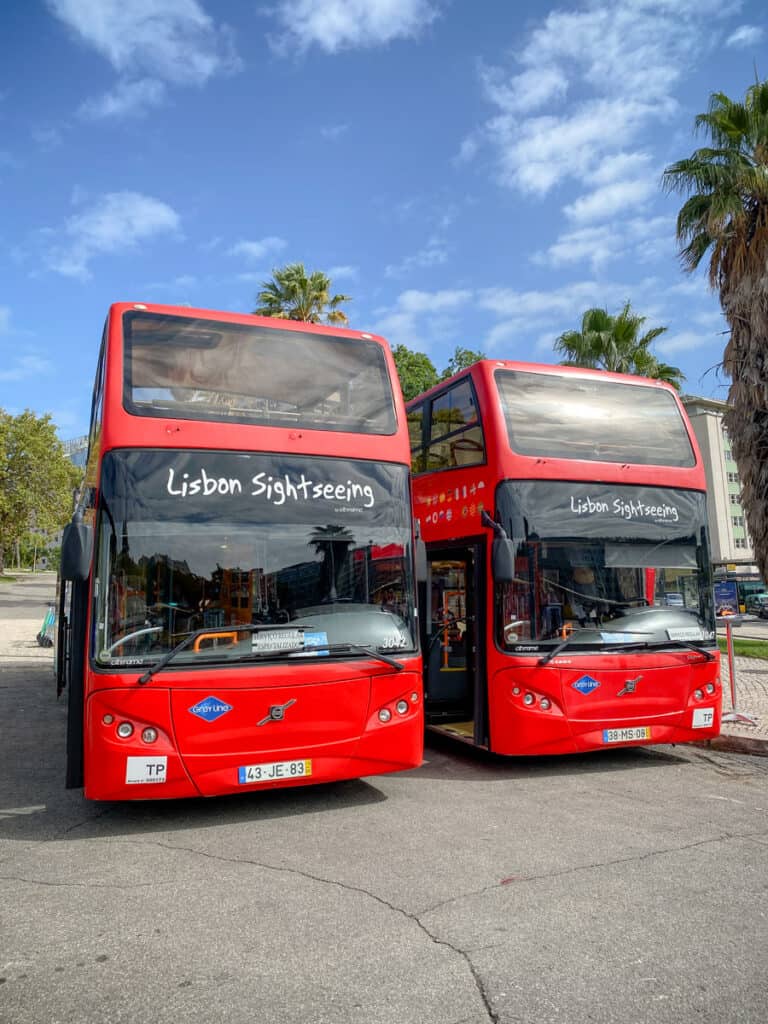 Two bright red double-decker buses parked side by side, both labeled "Lisbon Sightseeing" on the front, under a clear blue sky with scattered clouds. These hop-on hop-off Lisbon buses feature open tops and are ready for city tours, with palm trees and urban scenery in the background. Perfect for exploring Lisbon's landmarks.