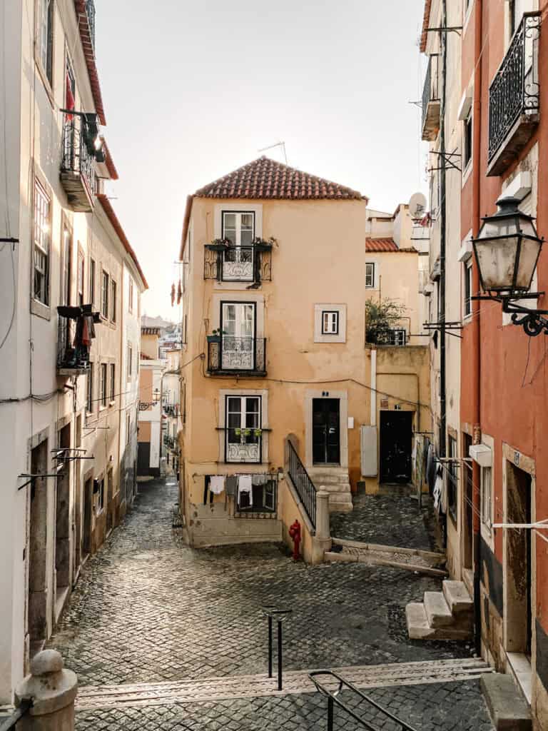 A sunlit cobbled street in Lisbon’s Alfama district with pastel buildings, wrought-iron balconies, and laundry hanging from lines. Ideal for those looking to learn photography in Lisbon with its charming, natural light and urban character.