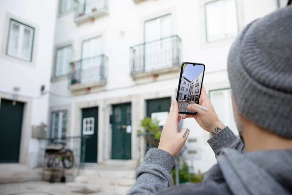A person in a grey hoodie photographs a white building with green doors using a smartphone, capturing the charm of Lisbon architecture. This image represents a hands-on photography tour in Lisbon.