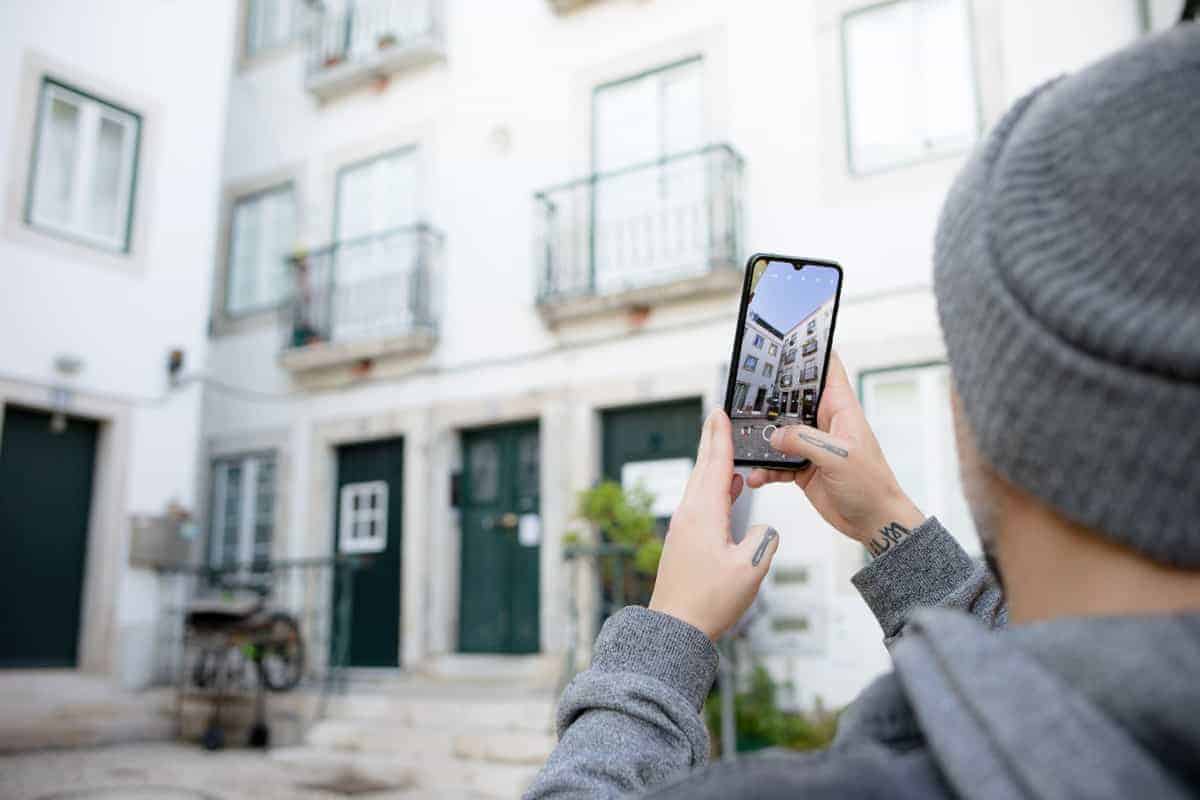 A person in a grey hoodie photographs a white building with green doors using a smartphone, capturing the charm of Lisbon architecture. This image represents a hands-on photography tour in Lisbon.