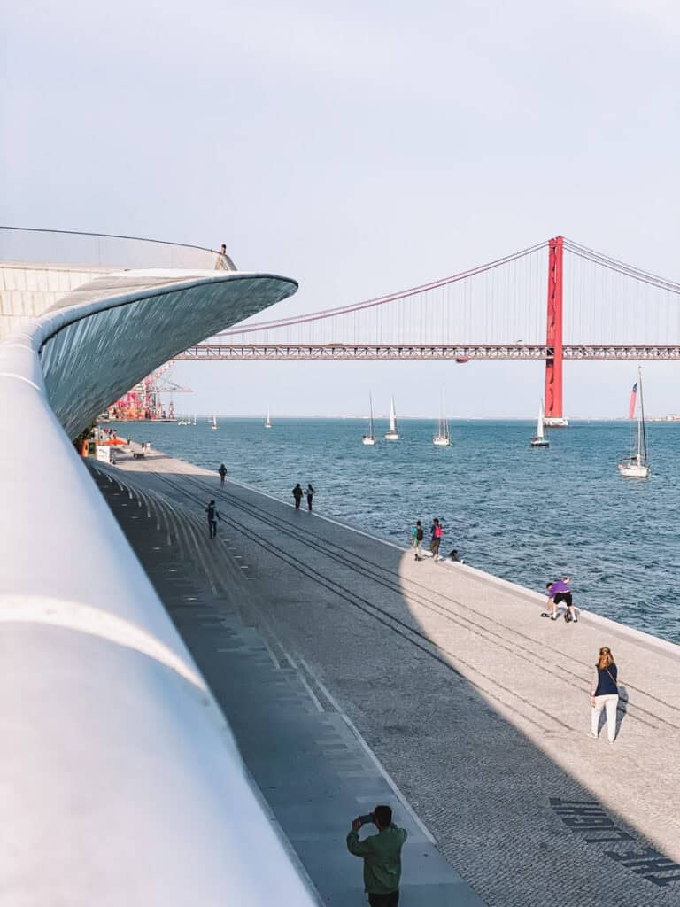 People stroll along the riverside promenade outside MAAT museum in Lisbon, with the red 25 de Abril Bridge in the background and sailboats on the Tagus River. A scenic setting for a photography class in Lisbon at MAAT.