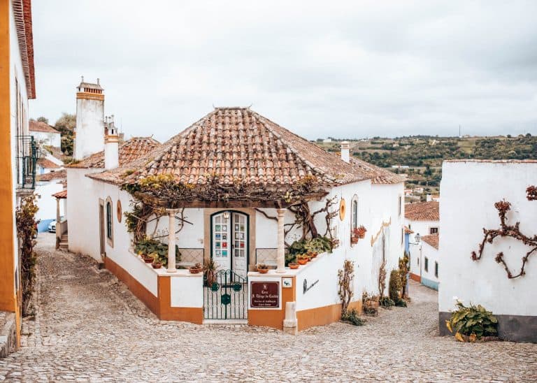 Een charmant wit huis met een betegeld dak en pergola in Óbidos, gelegen op een kruispunt van geplaveide straatjes met uitzicht op het groene landschap.
