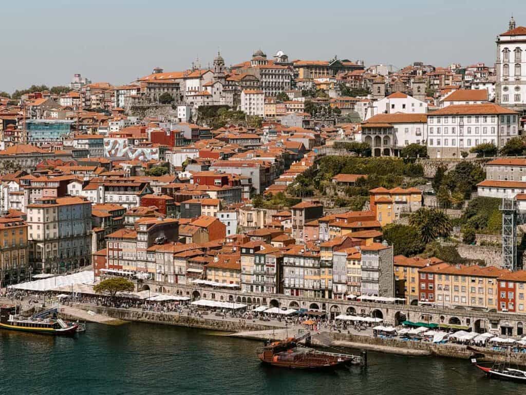 A panoramic view of Porto’s Ribeira district with colorful historic buildings lining the Douro River, bustling riverside cafés, and traditional Rabelo boats docked at the quay. This vibrant waterfront scene highlights one of the top things to do in Porto: exploring the UNESCO-listed old town and enjoying its riverside charm.