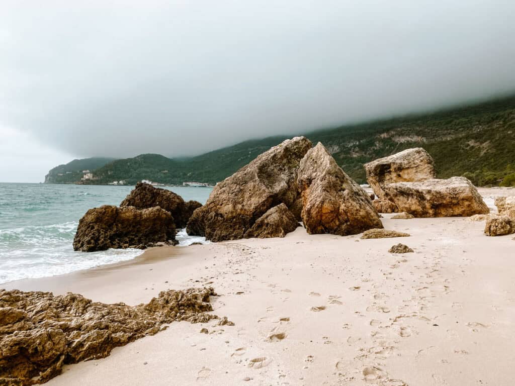 A moody coastal scene at Praia do Creiro, with large rock formations on the sandy shore and misty hills in the background.