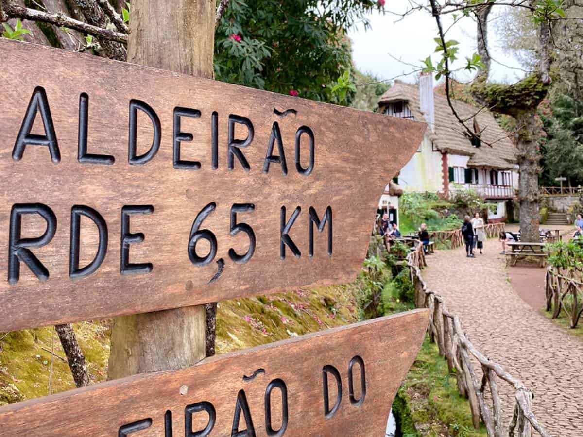 A close-up of a wooden trail sign points towards Aldeirão Verde, with a distance of 6.5 km carved into the wood. In the background, visitors stroll through a cobbled path beside traditional thatched-roof houses, capturing the charm of rural walks Madeira is known for.