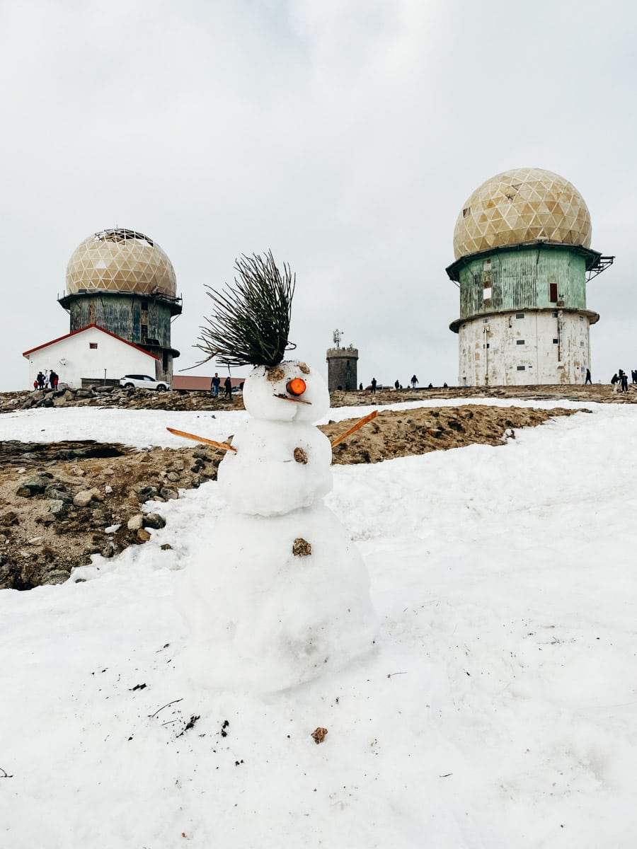 Een sneeuwpop met dennenappels, een wortelneus en takken als armen staat op een besneeuwd veld in Serra da Estrela, met op de achtergrond de karakteristieke koepelvormige radartorens.
