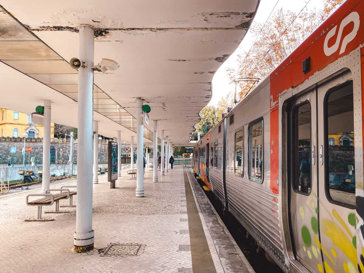Train at a platform on the Lisbon Sintra line, with graffiti-covered carriages and tiled flooring under a modern, curved canopy. A few passengers are visible in the distance, highlighting a typical scene of travel from Lisbon to Sintra by train.