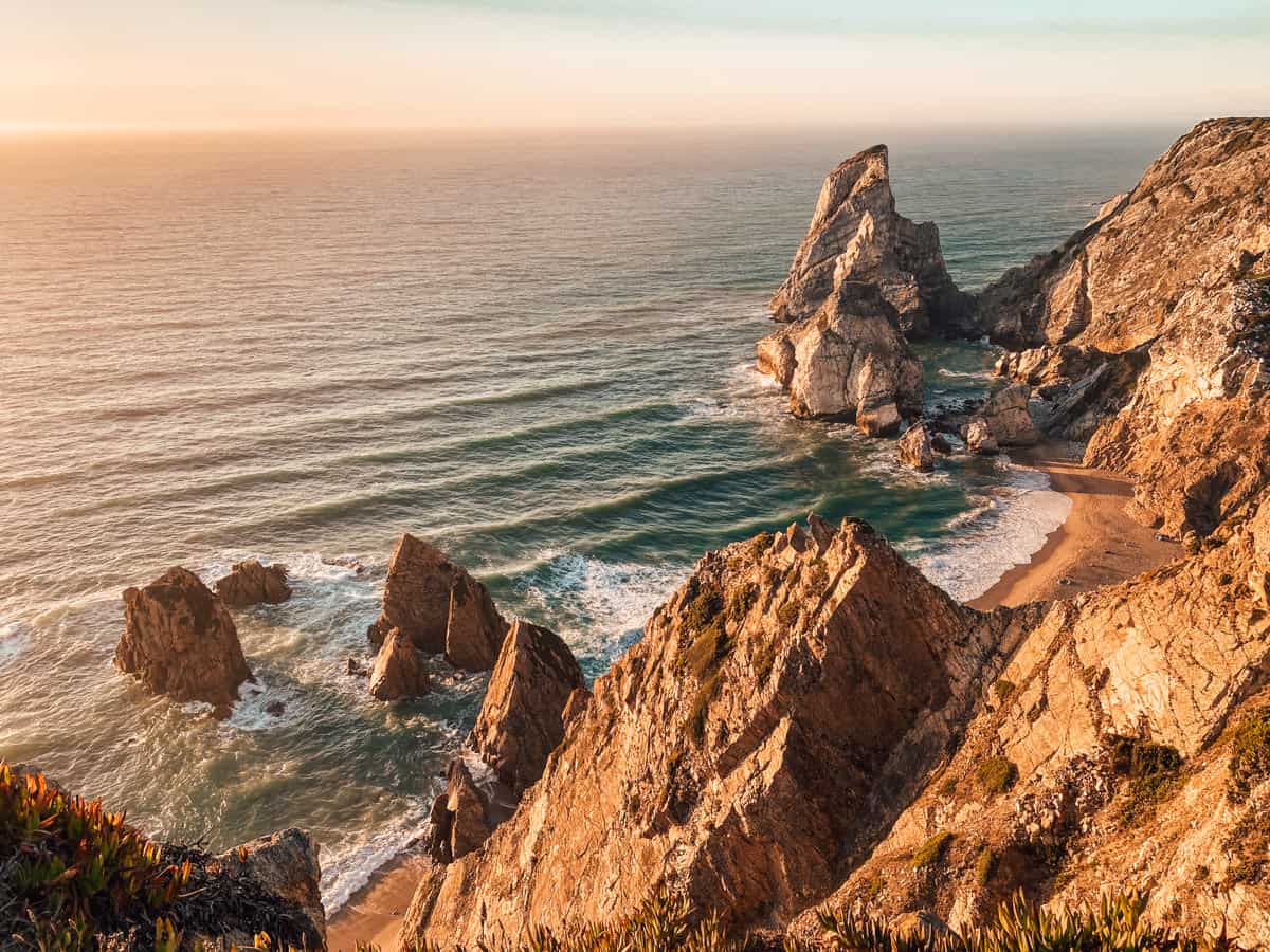 Praia da Ursa at sunset, near Cabo da Roca, Portugal, showcasing rugged cliffs descending to a secluded sandy beach. The golden light of the setting sun bathes the dramatic rock formations and tranquil ocean waves, creating a breathtaking coastal view.