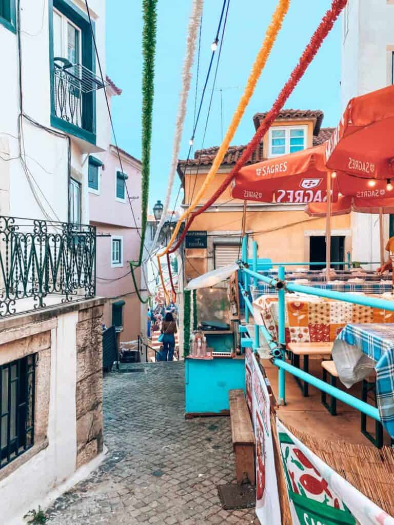 Smalle hellende straat in Alfama met kleurrijke slingers boven de terrassen tijdens Festas dos Santos Populares. Rode Sagres parasols en tafels met geruite kleden staan klaar voor feestvierders.