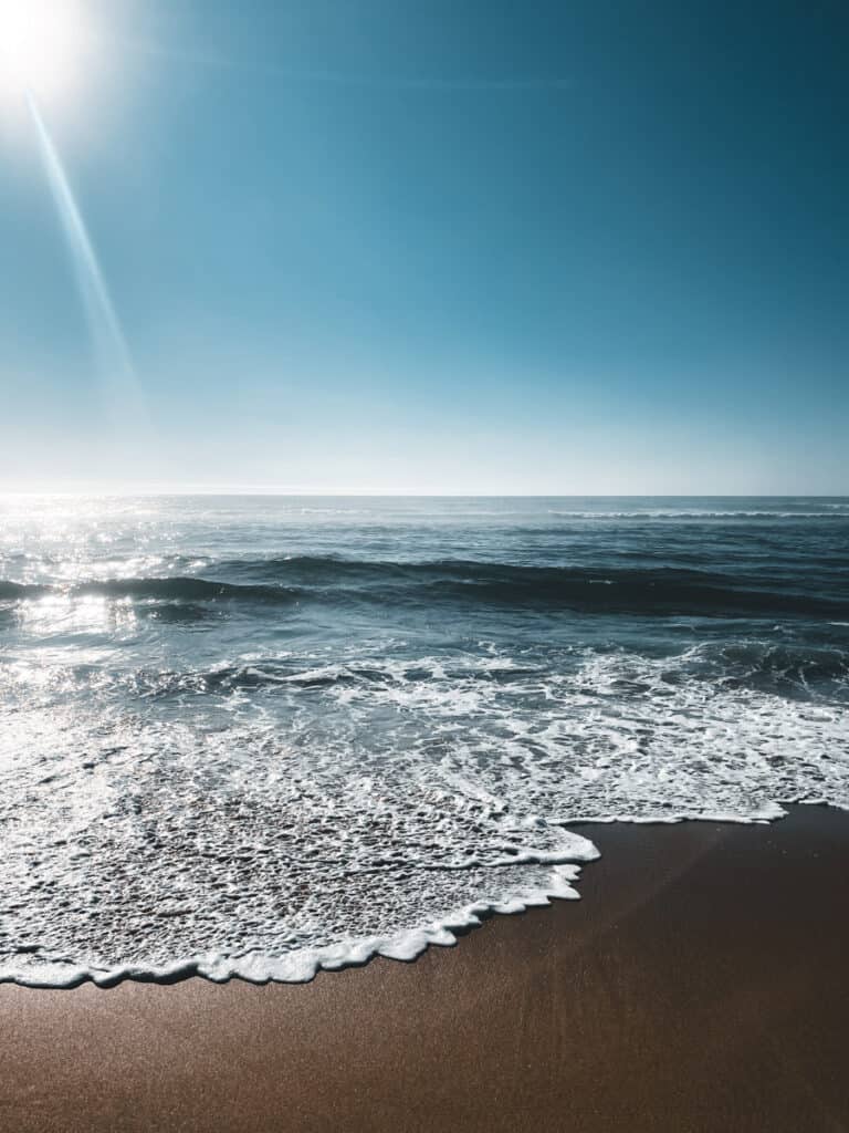 A sunlit morning at Praia Grande, a Sintra Beach in Portugal, with gentle waves washing up on the smooth sandy shore. The clear blue sky and sparkling Atlantic waters create a tranquil, inviting beach scene.