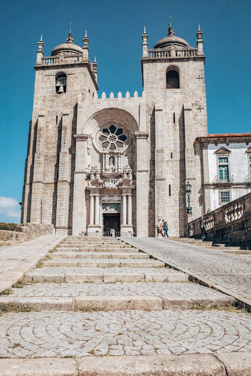 Trap van geplaveide stenen die omhoog leidt naar de ingang van de Kathedraal van Porto, geflankeerd door twee hoge klokkentorens met koepels, onder een heldere blauwe lucht.