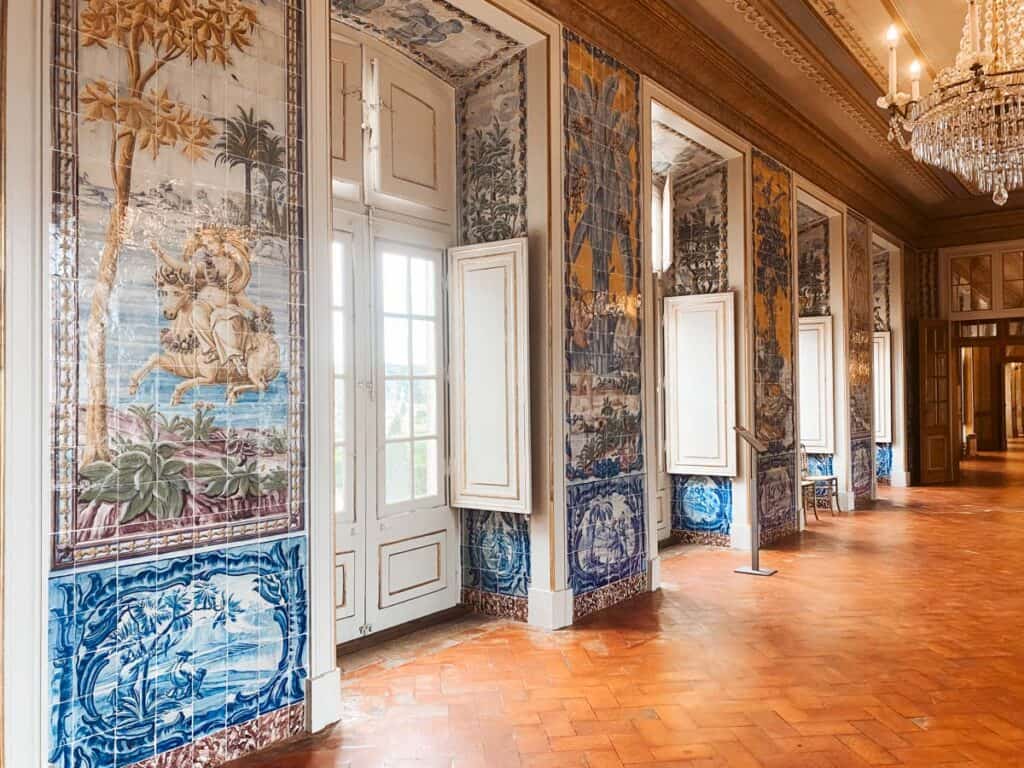 Interieur van Paleis Queluz met een lange gang versierd met kleurrijke azulejo-tegels die historische taferelen en landschappen afbeelden. Grote ramen met houten luiken, een glanzende parketvloer en een kristallen kroonluchter benadrukken de elegante barokstijl van het paleis.