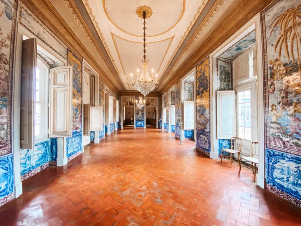 De Corridor of Sleeves in Paleis Queluz, een lange gang met terracottakleurige vloer, witte houten raamluiken en kleurrijke azulejo-tegels met levendige scènes en landschappen. Een kristallen kroonluchter hangt centraal, en enkele elegante stoelen staan langs de muur, wat de grandeur van de ruimte benadrukt.