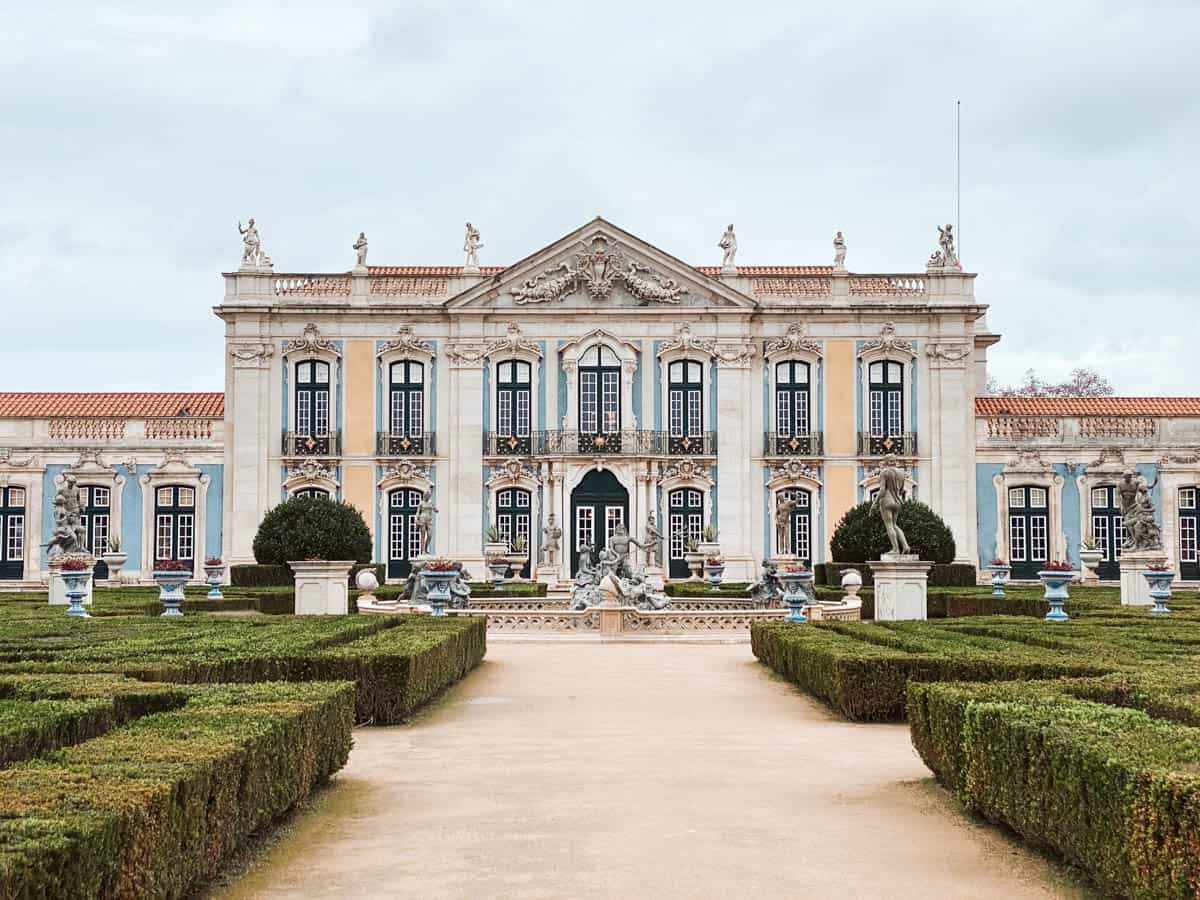 De symmetrische voorgevel van het Nationaal Paleis van Queluz met pastelkleurige muren, sierlijke balkons en beelden op het dak. Voor het paleis liggen perfect gesnoeide tuinen met standbeelden, bloempotten en een centrale fontein.