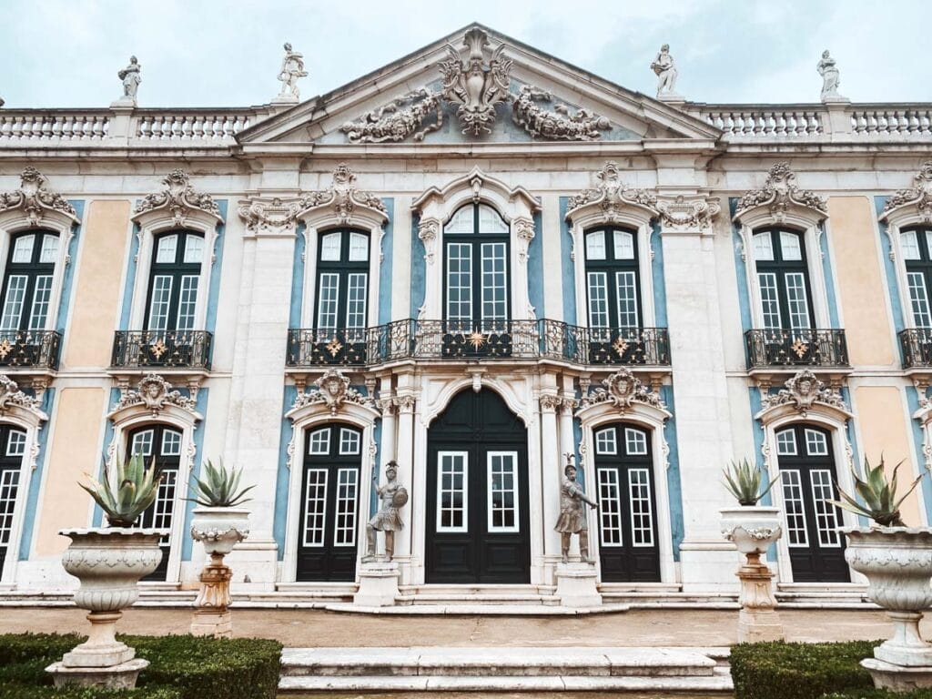 De voorgevel van het Nationaal Paleis van Queluz, een sierlijk gebouw met barokke architectuur, hoge ramen met zwarte luiken en sierlijke balkons. Beeldhouwwerken en grote planten in stenen potten flankeren de ingang, wat bijdraagt aan de koninklijke uitstraling van het paleis.