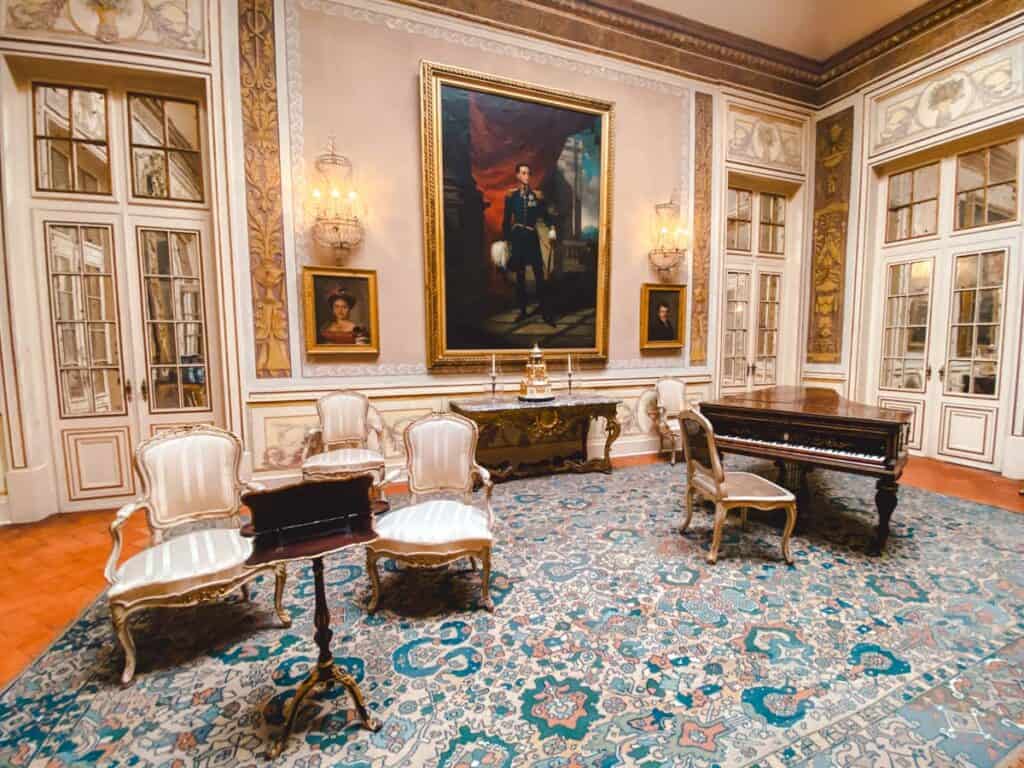 De Lantaarnzaal van Paleis Queluz met een rijkversierde inrichting, waaronder een houten piano, elegante witte stoelen, een sierlijke tafel en een groot vloerkleed met blauw patroon. Aan de muur hangen portretten, waaronder een imposant schilderij van een man in militair uniform, omlijst door kristallen wandkandelaars.