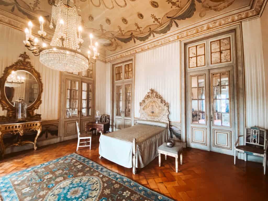 Koninklijke slaapkamer in Paleis Queluz, ooit de vertrekken van prinses Maria Francisca Benedita, met een elegant bed met sierlijk hoofdbord, een grote kristallen kroonluchter en rijk versierde spiegels en meubels. De ruimte straalt zachte luxe uit met pastelkleuren, gouden accenten en een gedecoreerd plafond.