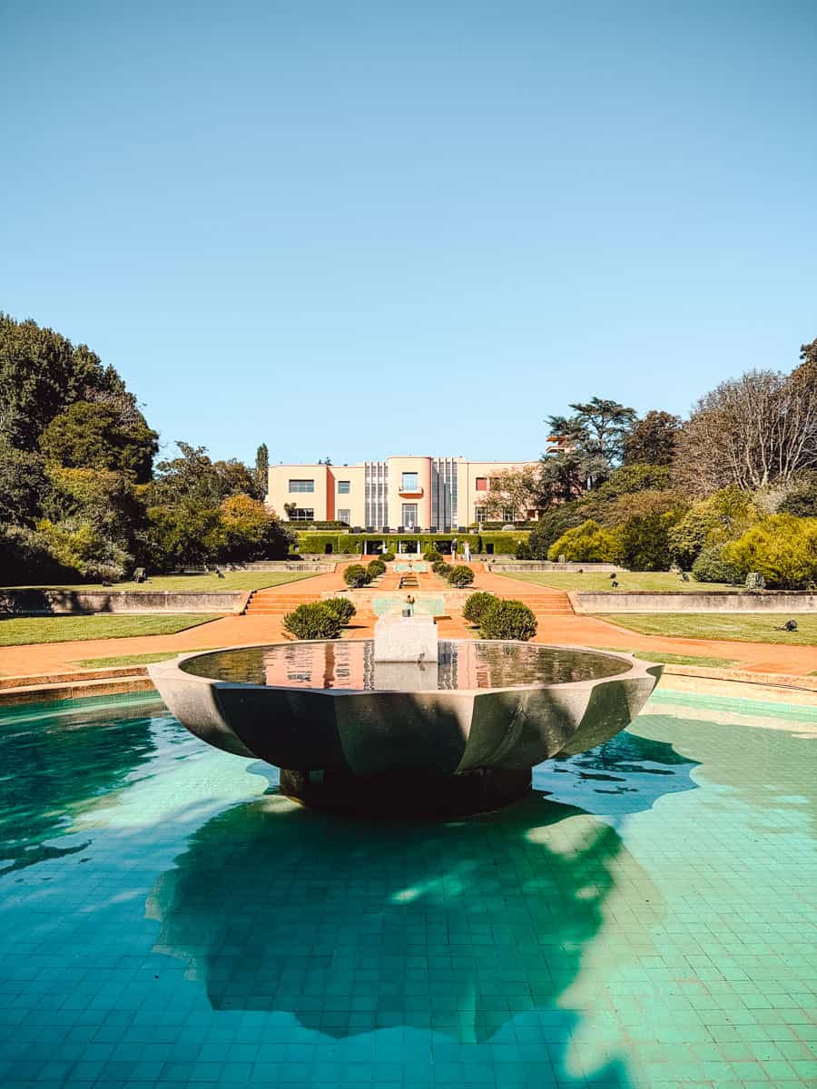 Een prachtige blik op de Serralves villa in Porto, omringd door groene tuinen en een serene waterpartij. Op de voorgrond weerspiegelt een moderne fontein de blauwe lucht, terwijl het art-deco herenhuis in de achtergrond de aandacht trekt. De Serralves villa is een iconisch cultureel en architectonisch hoogtepunt in Portugal.