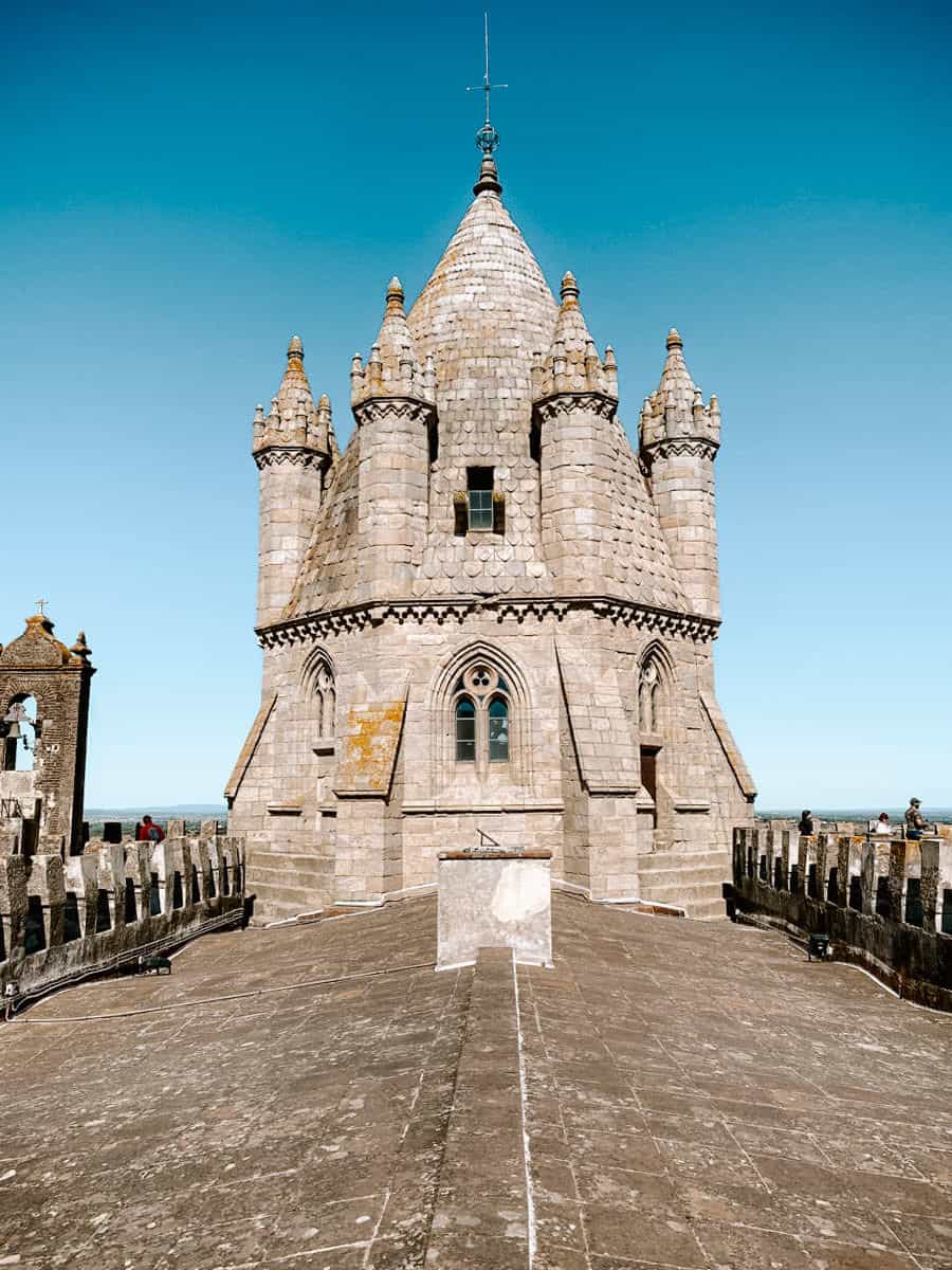 De indrukwekkende kathedraal van Évora met zijn gotische torens en stenen details, tegen een heldere blauwe lucht, met een brede loopbrug op het dak die een panoramisch uitzicht over de omgeving biedt.