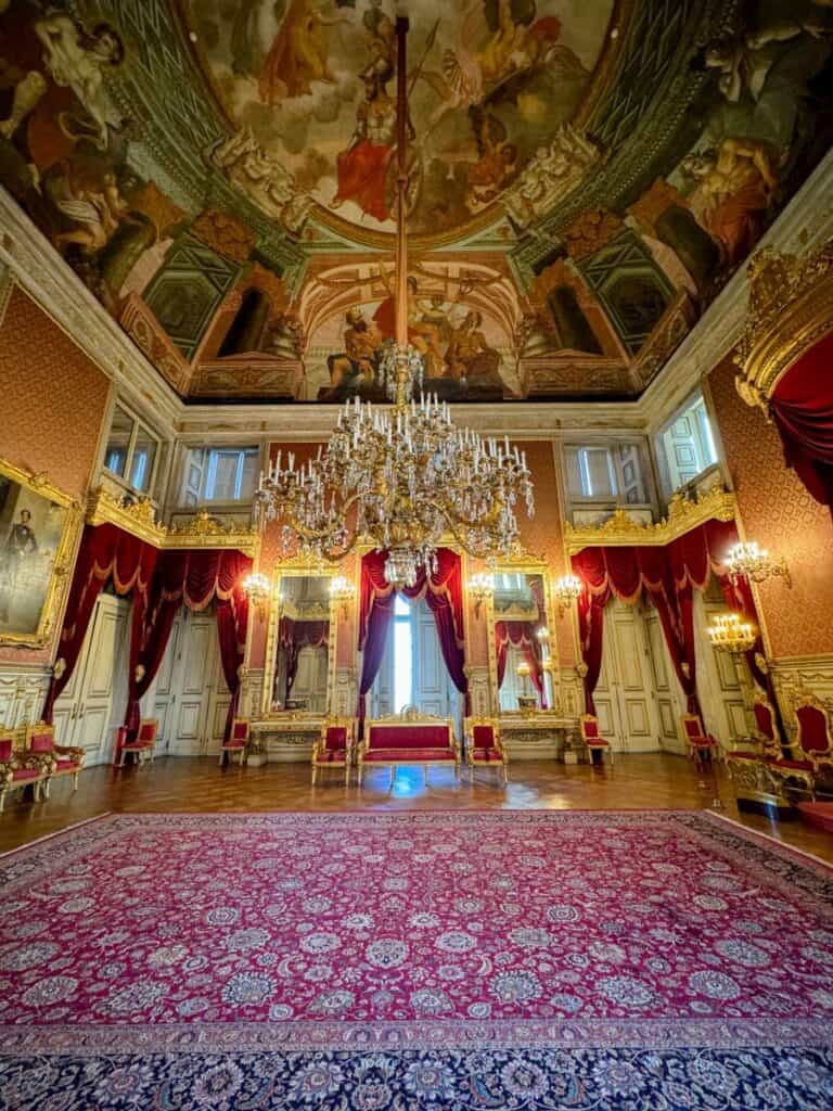 De luxueuze troonzaal van het Ajuda Paleis in Lissabon, versierd met een imposante kristallen kroonluchter, rode fluwelen gordijnen en vergulde spiegels. De rijkelijk gedecoreerde plafondfresco’s tonen mythologische taferelen, terwijl de koninklijke meubels en het grote tapijt de grandeur van het paleis benadrukken. Dit voormalige koninklijke verblijf weerspiegelt de weelderige Portugese architectuur en geschiedenis.