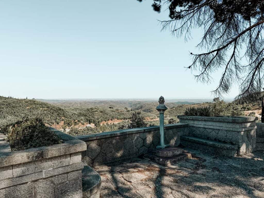 A peaceful viewpoint at Caldas de Monchique, Portugal, offering a panoramic view of rolling green hills stretching towards the horizon. The stone terrace features a rustic water fountain and built-in benches, shaded by overhanging tree branches, creating a serene and picturesque resting spot in the Monchique mountains.