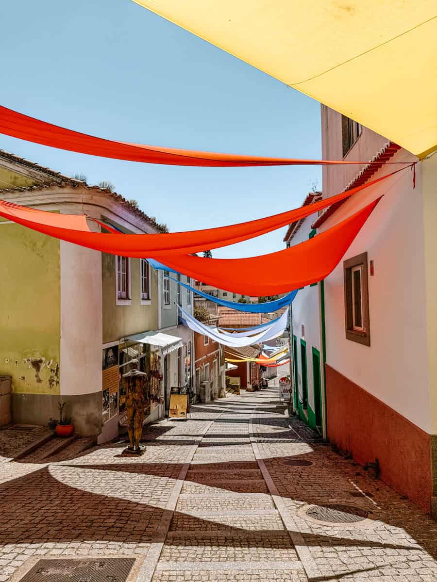 Een sfeervol straatje in Monchique, Algarve, met kleurrijke zonneschermen in rood, geel en blauw die schaduw werpen over de geplaveide weg. De charmante pastelkleurige gebouwen langs de straat huisvesten lokale winkels en cafés, wat de authentieke Portugese sfeer benadrukt.
