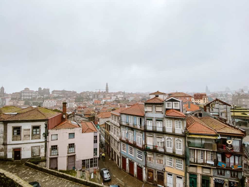 Uitzicht bij de Porto Kathedraal over smalle straten en oude huizen met rode dakpannen. De skyline van Porto ligt in de mistige achtergrond onder een grijze regenlucht.