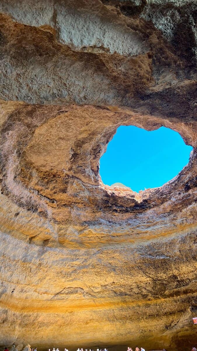 Uitzicht van binnenuit op de grote natuurlijke opening in het plafond van de Benagil Caves, met blauwe lucht zichtbaar door het gat