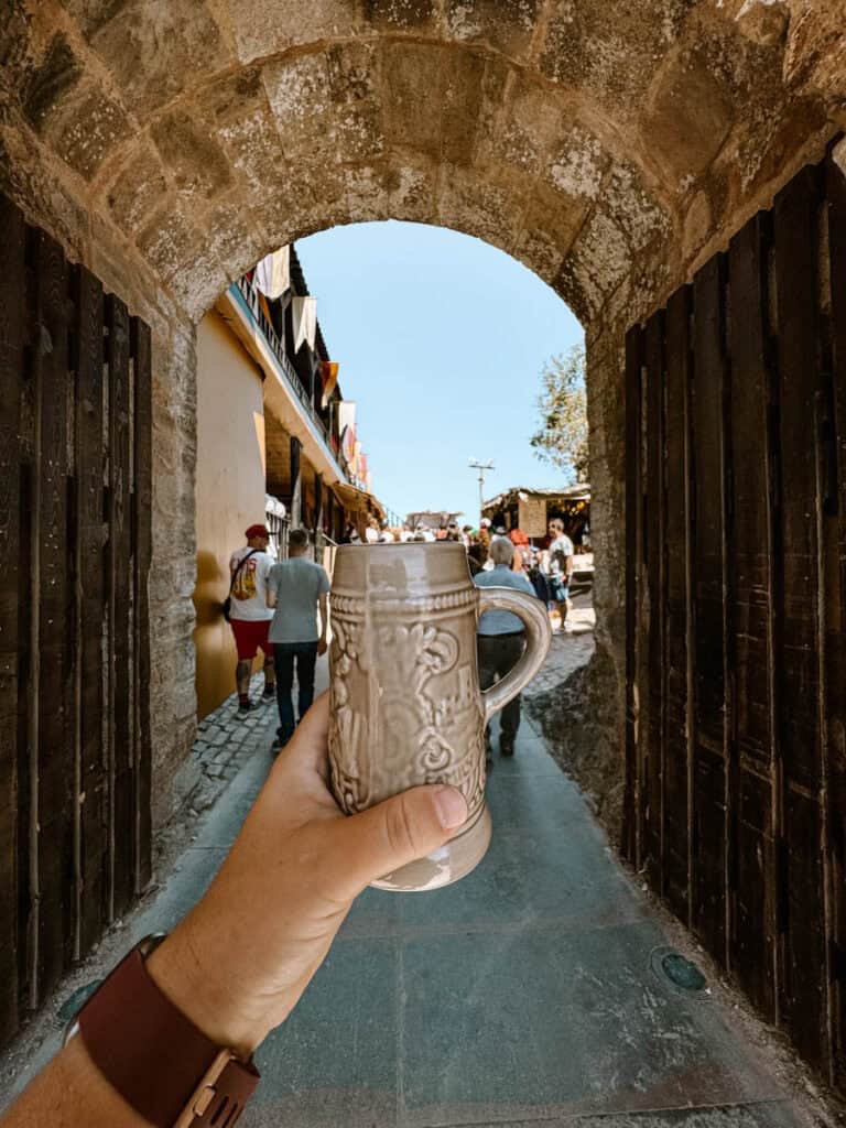 A ceramic tankard held up under a stone archway at the Mercado Medieval Óbidos, with festival-goers and medieval-style buildings visible in the background. The stein features intricate relief patterns, adding to the historic feel of the scene.
