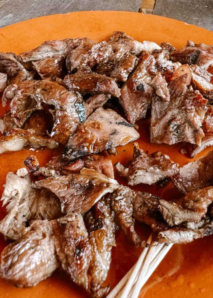 A terracotta plate filled with grilled pork slices at Mercado Medieval Óbidos, served juicy and charred, with toothpicks provided for easy sampling. The dish reflects rustic medieval-style Portuguese cooking.