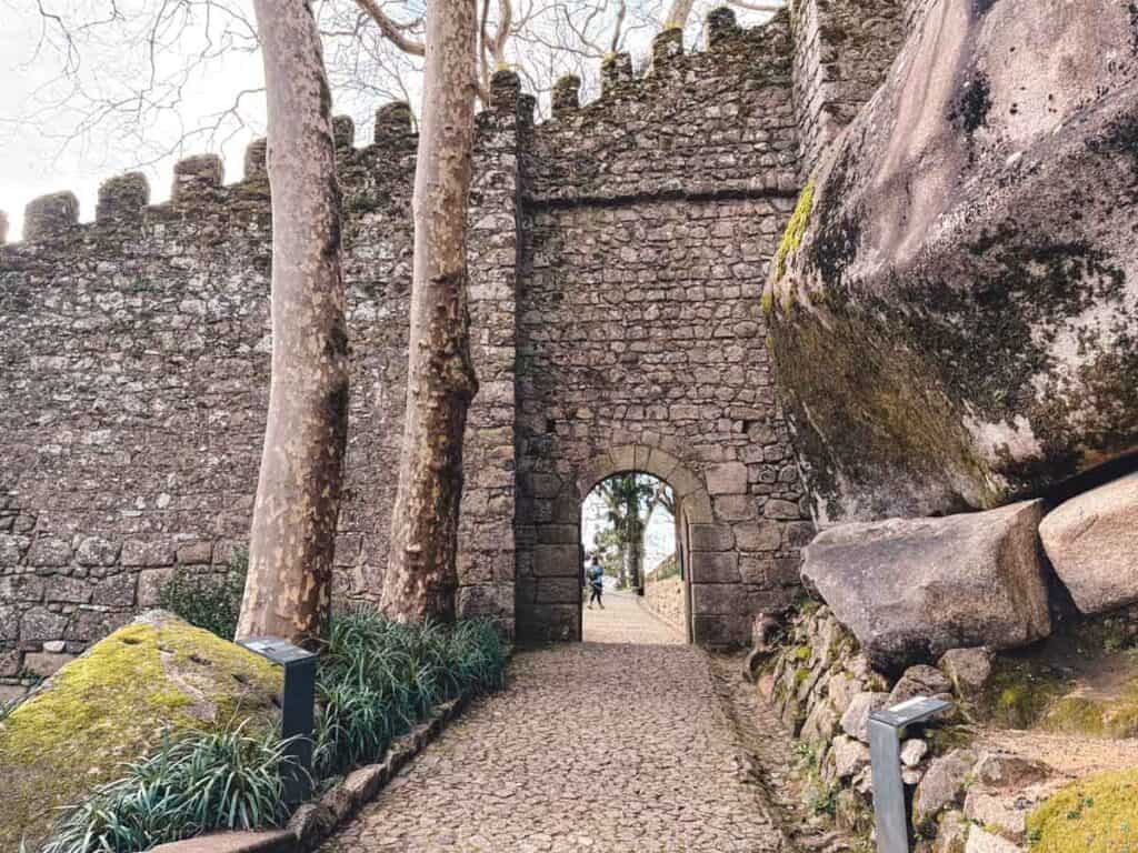 Ingangspoort van Castelo dos Mouros met een geplaveid pad, geflankeerd door eeuwenoude stenen muren, bomen en grote met mos bedekte rotsen. De middeleeuwse architectuur en natuurlijke elementen versterken de historische sfeer van het kasteel in Sintra.