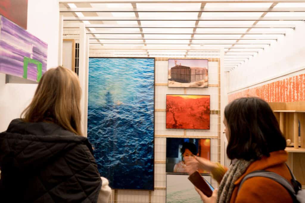 Two visitors admire a vivid selection of contemporary artworks in a Lisbon art gallery, including a striking large photograph of the ocean, vibrant desert landscapes, and a portrait. The well-lit display area is narrow and modern, filled with visual contrast and colour.