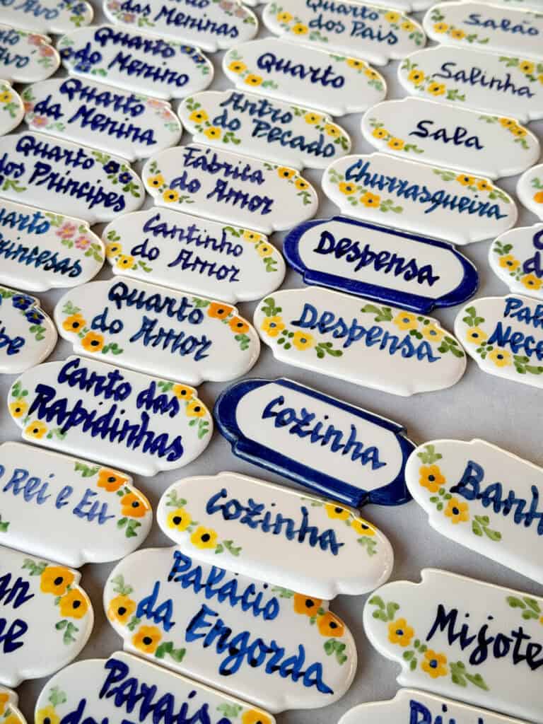Placas de cerâmica pintadas à mão à venda na Feira da Ladra com flores amarelas e letras azuis para identificar divisões da casa. Entre os textos visíveis estão "Quarto das Meninas" "Quarto dos Pais" "Salão" "Quarto do Menino" "Quarto da Menina" "Quarto dos Príncipes" "Sítio do Pecado" "Fábrica do Amor" "Cantinho do Amor" "Sala" "Salinha" "Churrasqueira" "Despensa" "Quarto do Amor" "Cozinha" e "Palácio da Engorda".