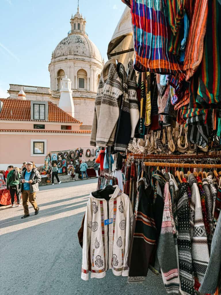 Banca de roupas vintage em Lisboa com casacos de lã e peças coloridas penduradas em primeiro plano. Pessoas caminham entre as barracas com a cúpula do Panteão Nacional ao fundo.
