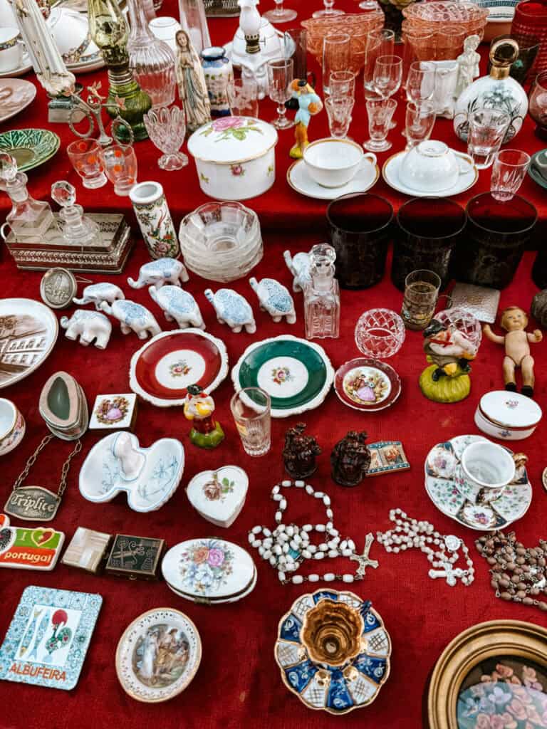 Banca de antiguidades em um dos mercados em Lisboa com louças de porcelana copos de cristal pequenas esculturas frascos decorativos e rosários sobre uma toalha vermelha. Entre as lembranças aparecem as inscrições “I ♥ portugal” e “ALBUFEIRA”.