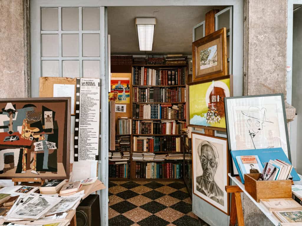 Livraria e banca de arte em uma feira de antiguidades em Lisboa com estantes cheias de livros antigos e mesas cobertas por gravuras retratos e obras emolduradas. Entre os textos legíveis aparecem “LA SCUOLA AL CINEMA” e “FERNANDO PESSOA” em meio a livros cartazes e ilustrações expostos na entrada.