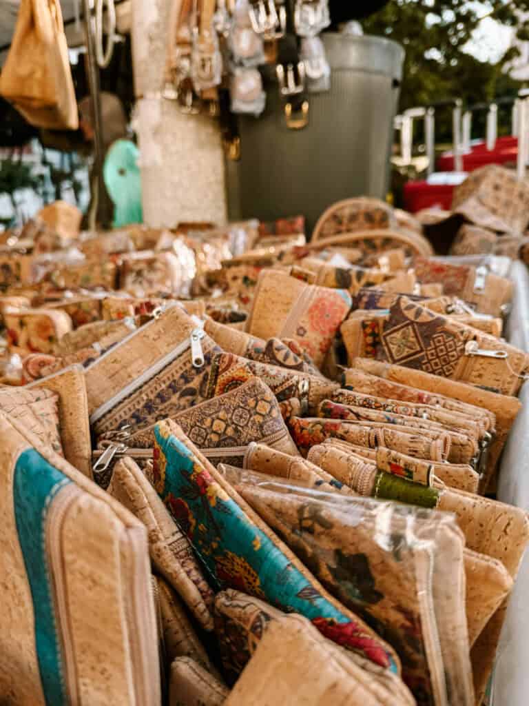 Carteiras bolsas e pequenos acessórios de cortiça com diferentes estampas estão organizados em uma banca de mercado de pulgas em Lisboa. Os produtos aparecem em tons naturais de bege com detalhes coloridos e zíperes metálicos.