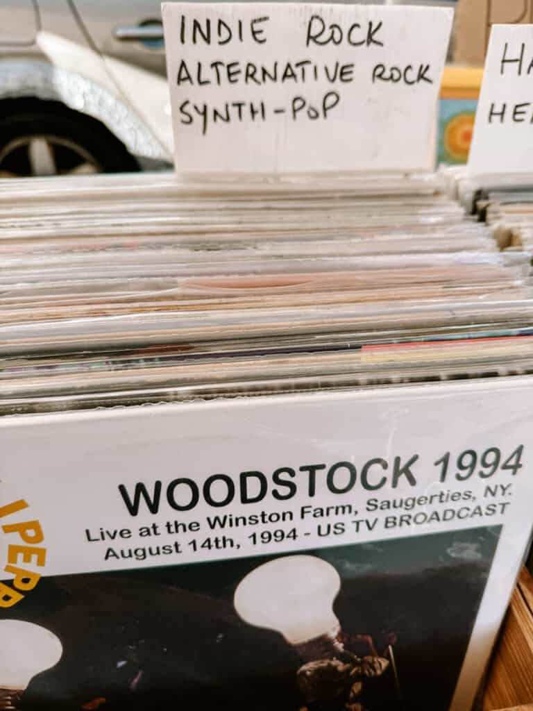 Discos de vinil organizados em caixas em uma feira de vinil no Mercado de Santa Clara com uma placa escrita "INDIE ROCK" "ALTERNATIVE ROCK" e "SYNTH-POP". Em primeiro plano aparece um disco com os textos "WOODSTOCK 1994" "Live at the Winston Farm, Saugerties, NY" e "August 14th, 1994 - US TV BROADCAST".