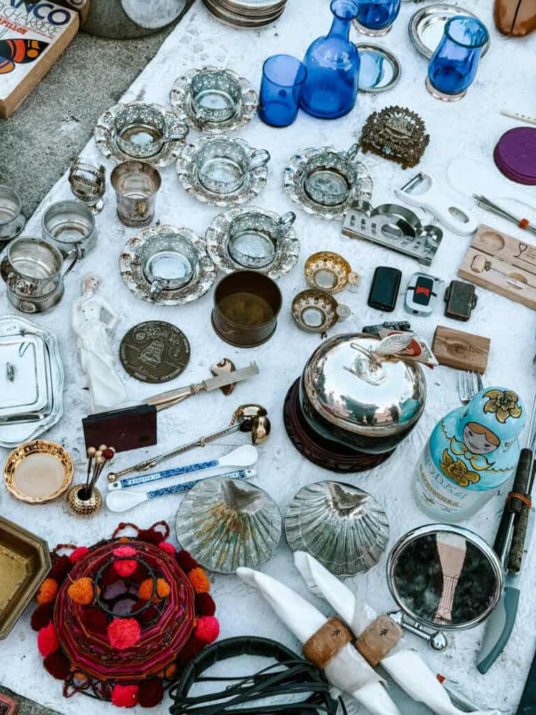 Objetos vintage e antiguidades à venda em uma banca com xícaras de vidro garrafas azuis peças de metal louças espelhos e pequenos itens decorativos. A variedade de peças usadas mostra o tipo de achado que pode aparecer durante compras em Lisboa.