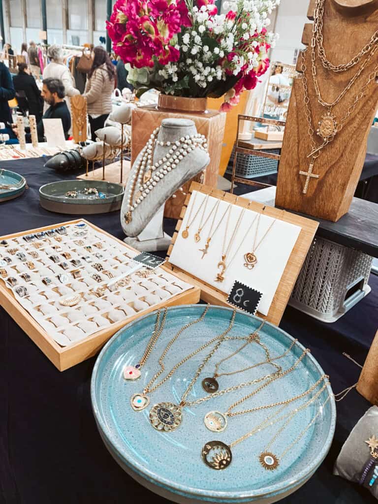 Banca de joias em um mercado de rua em Lisboa com anéis colares dourados pérolas e pingentes expostos sobre bandejas e suportes de madeira. Um vaso com flores cor de rosa e brancas decora a mesa enquanto visitantes circulam ao fundo.