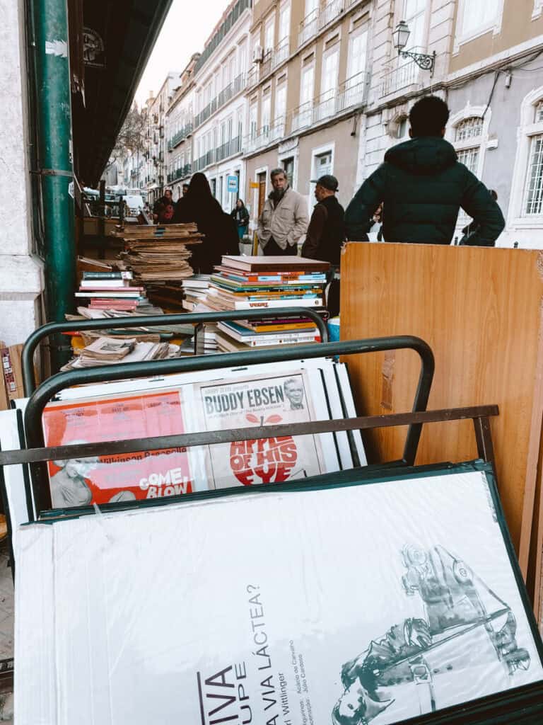 Banca de livros discos cartazes e impressos usados espalhados pela calçada em uma cena de compras em Lisboa. Pessoas conversam junto às pilhas de livros e entre os títulos visíveis estão “BUDDY EBSEN” e “COME BLOW YOUR HORN”.