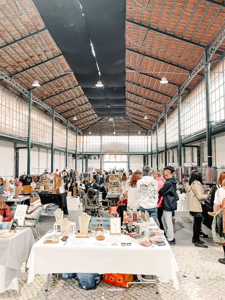 Interior do Mercado de Santa Clara com bancas de joias artesanato roupas e acessórios distribuídas sob uma estrutura alta de ferro e madeira. Visitantes circulam entre as mesas e uma pessoa ao centro usa um casaco com o texto "STAR WARS".