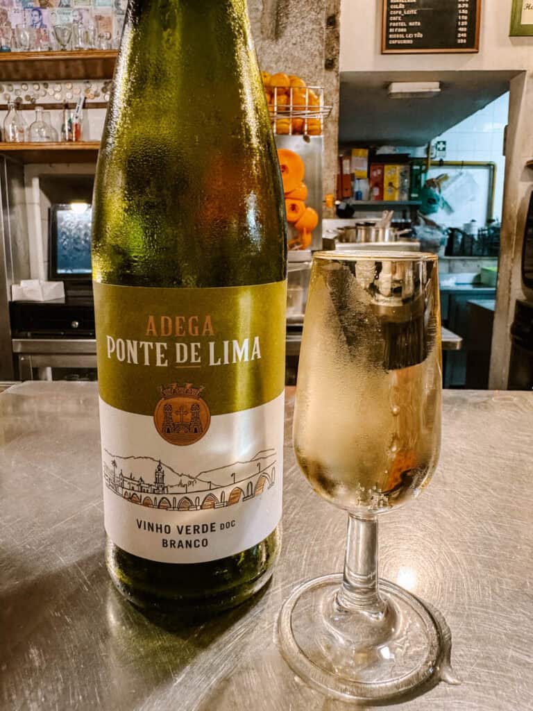 Garrafa de vinho verde branco "Adega Ponte de Lima" ao lado de uma taça servida num balcão de restaurante. Provar vinhos portugueses num espaço interior é uma experiência acolhedora para aproveitar Lisboa no inverno.