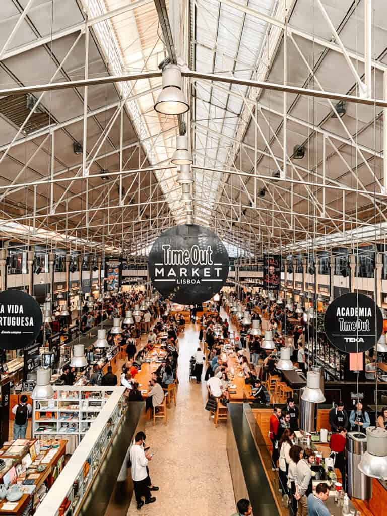 Interior movimentado do Time Out Market Lisboa com longas mesas partilhadas bancas de comida e dezenas de visitantes sob uma grande cobertura metálica. O mercado coberto é uma opção popular de passeios cobertos em Lisboa para comer provar especialidades locais e passar algumas horas longe da chuva.