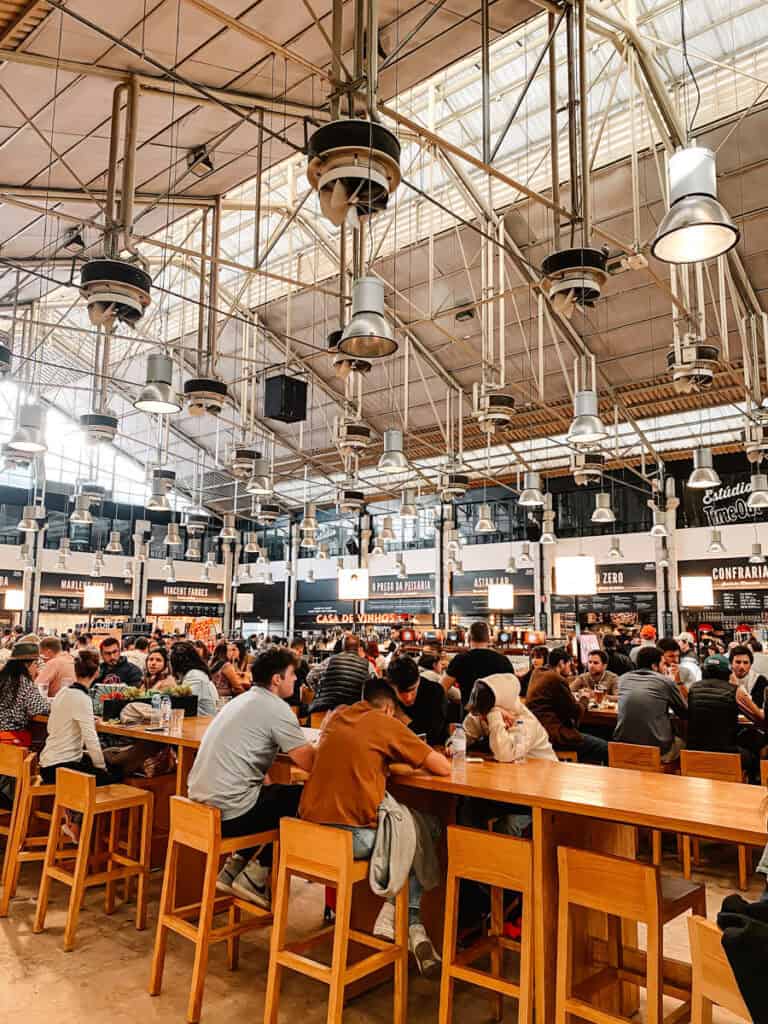 Mesas de madeira cheias de visitantes no interior do Time Out Market Lisboa com várias bancas de comida ao fundo e grandes candeeiros suspensos na estrutura metálica. Comer e experimentar diferentes sabores num mercado coberto é uma das atividades em Lisboa com chuva.
