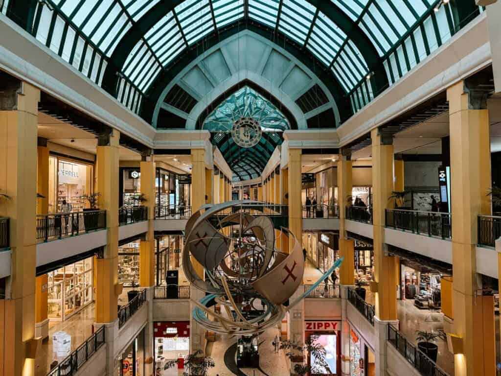 Interior do centro comercial colombo em Lisboa com vários pisos de lojas sob uma grande cobertura de vidro e uma escultura inspirada na navegação ao centro. Entre as montras visíveis aparecem "MERRELL" "ZIPPY" e "GUESS" num espaço coberto para fazer compras em Lisboa.