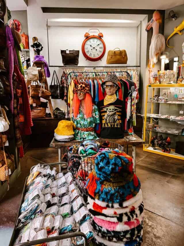 Loja vintage, às de espadas, em Lisboa cheia de roupas coloridas chapéus acessórios e objetos decorativos expostos em cabides prateleiras e cestos. Explorar lojas cobertas como esta é uma das atividades em Lisboa com chuva para descobrir peças diferentes sem depender do tempo.