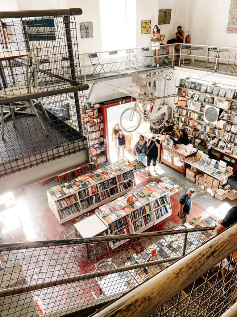 Interior da livraria Ler Devagar em Lisboa com estantes cheias de livros mesas em dois pisos e uma bicicleta suspensa como parte da decoração. A livraria é uma paragem acolhedora para aproveitar Lisboa em dia de chuva enquanto se explora um dos espaços culturais mais conhecidos da cidade.